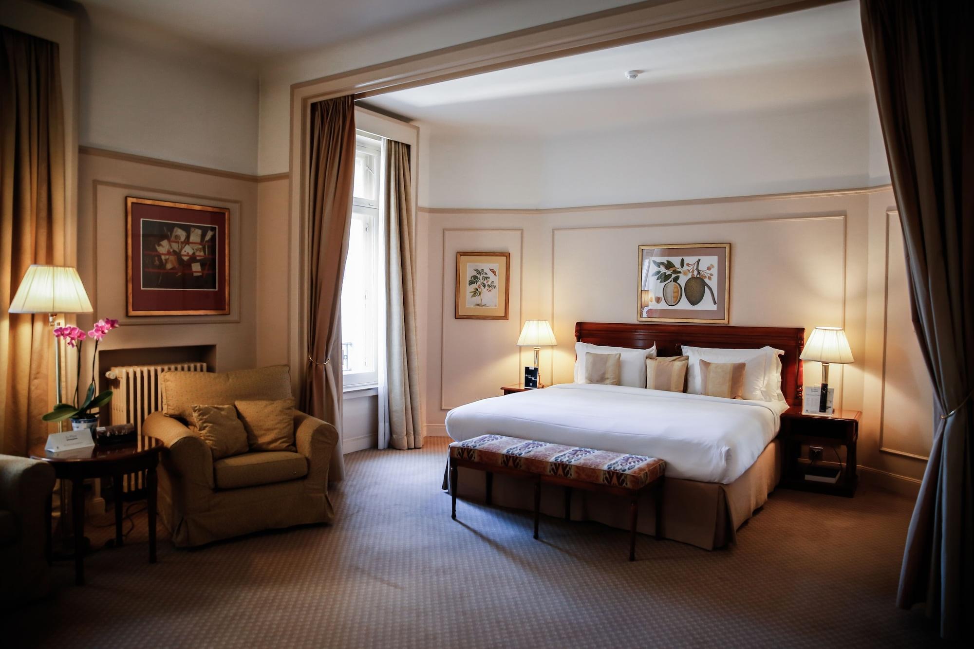 Hotel Le Plaza Brusel Exteriér fotografie The photo shows a well-decorated bedroom. It features a large bed with white linens and several pillows, positioned against a wall with framed artwork above it. On either side of the bed, there are lamps providing soft lighting. There’s a cozy seatin