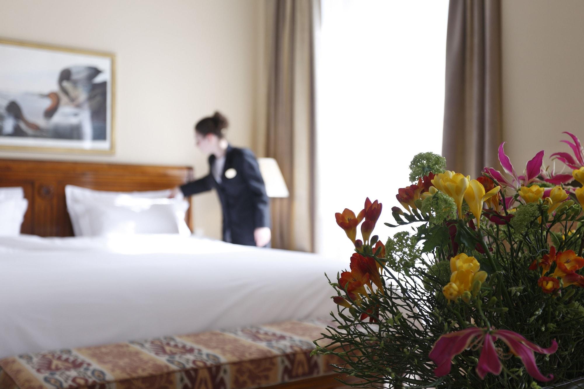 Hotel Le Plaza Brusel Exteriér fotografie The photo shows a hotel room with a neatly made bed in the background. A staff member, dressed in a uniform, is fixing the bed. In the foreground, there is a bouquet of colorful flowers, adding a touch of vibrancy to the scene. The room is well-lit, 