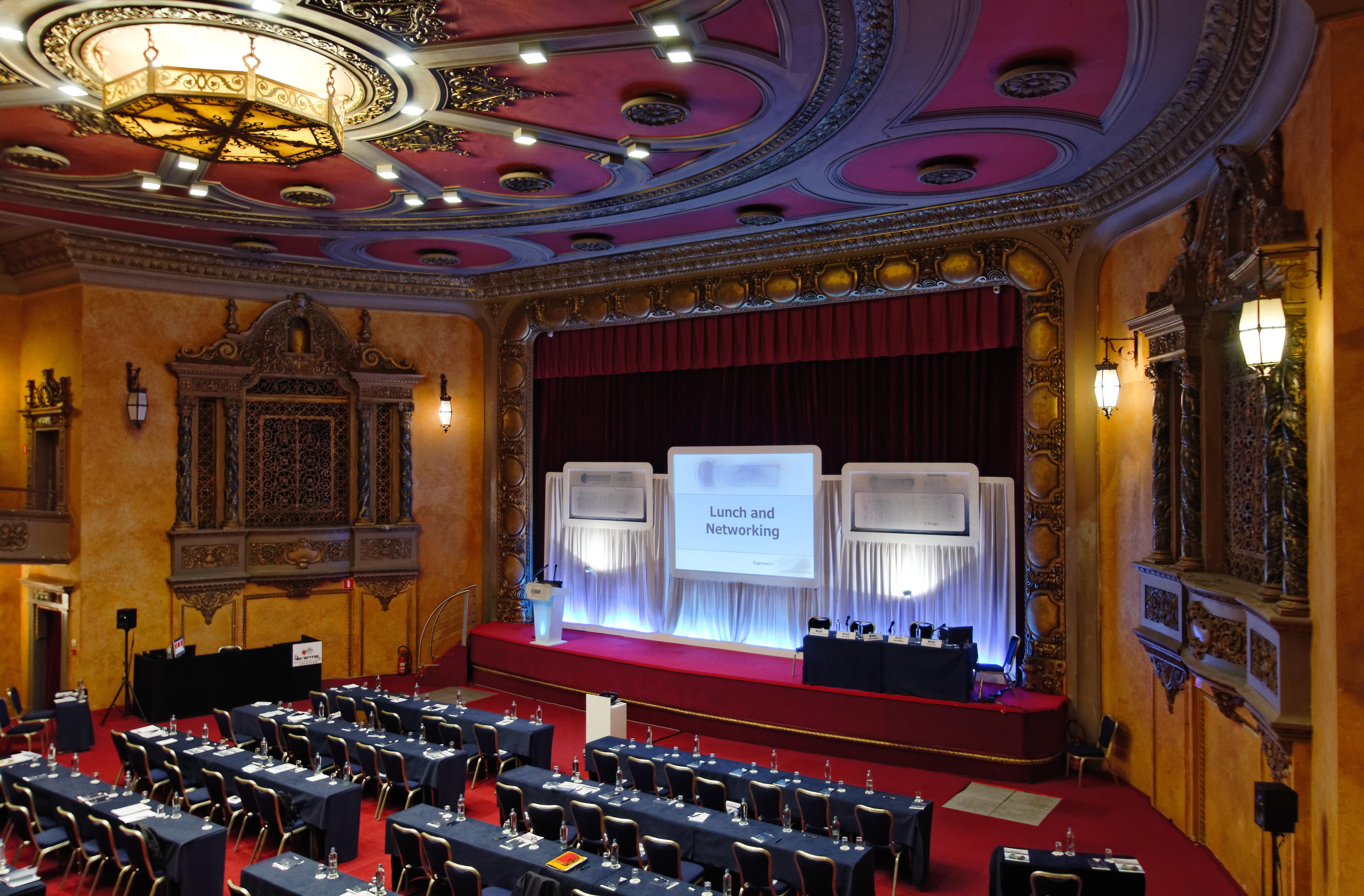 Hotel Le Plaza Brusel Exteriér fotografie The photo shows a spacious banquet or conference hall with a stage set up for an event. The stage features a large screen with the words "Lunch and Networking" displayed prominently. In front of the stage, there are several rows of neatly arranged ta