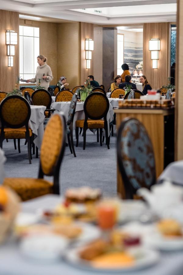Hotel Le Plaza Brusel Exteriér fotografie The photo depicts an elegant restaurant interior. There are several round tables covered with white tablecloths, each accompanied by ornate chairs featuring a gold and dark blue design. In the background, various groups of people are seated at differ