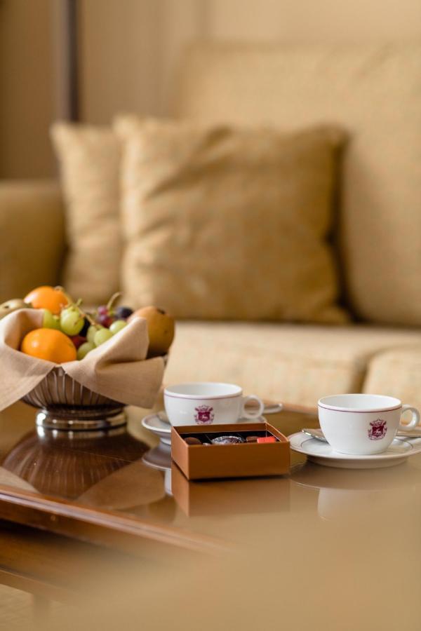 Hotel Le Plaza Brusel Exteriér fotografie The photo shows a cozy setting with two small white cups placed on saucers, likely for coffee or tea. Next to the cups, there is a small box of chocolates. In the foreground, a silver bowl holds a variety of fruits, including oranges, grapes, and oth