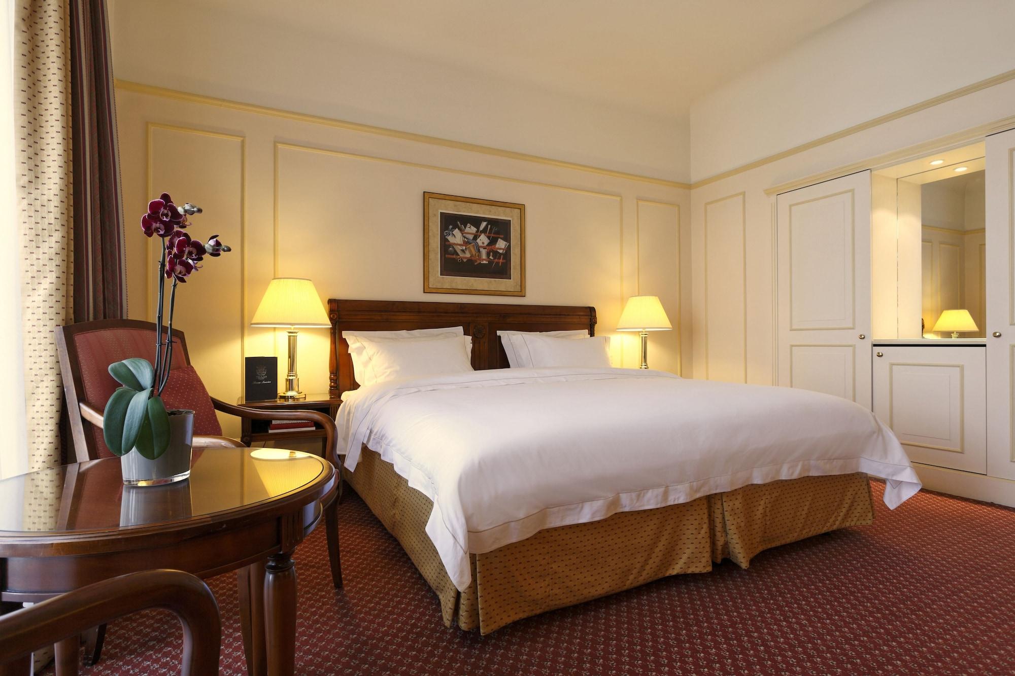 Hotel Le Plaza Brusel Exteriér fotografie The photo shows a cozy hotel room featuring a large bed with white linens and a golden bed skirt. On either side of the bed, there are bedside lamps providing warm light. There's a small table with a vase containing purple orchids, adding a touch of 