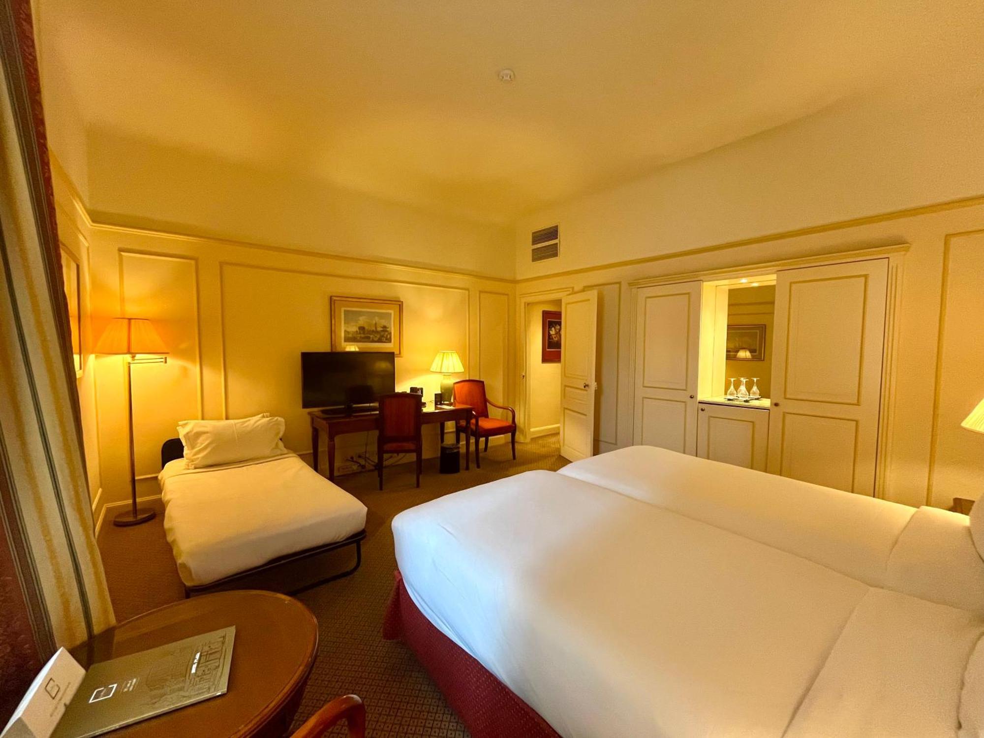 Hotel Le Plaza Brusel Pokoj fotografie The photo shows an interior of a hotel room with a warm, cozy ambiance. It features two single beds made with white linens positioned side by side. There is a small, rolled-up cot or mattress in the corner. Near the beds, there is a bedside table and