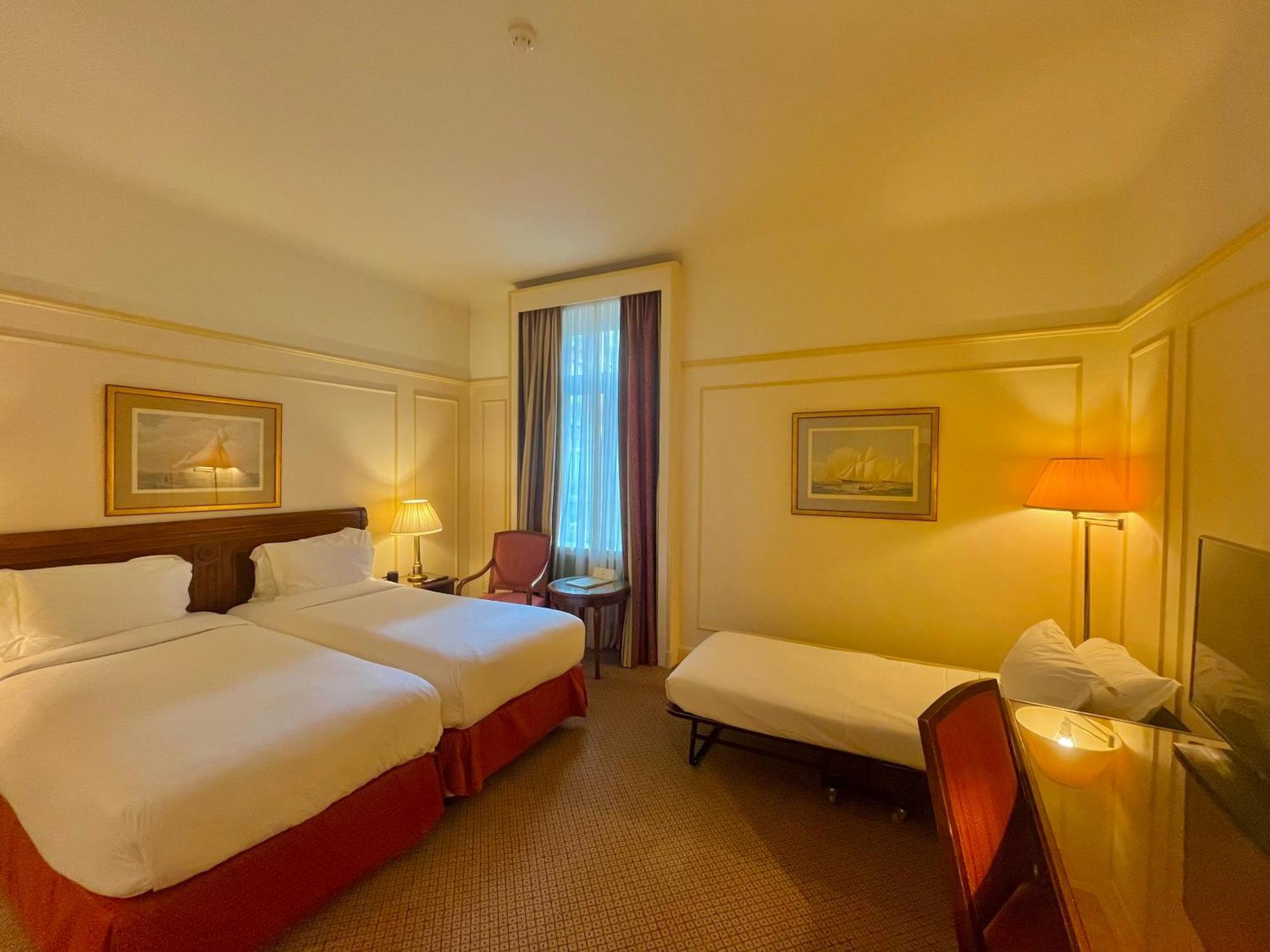 Hotel Le Plaza Brusel Pokoj fotografie The photo shows a hotel room featuring two single beds with white linens and red accents. There is a small extra bed on the floor, along with a wooden chair. A desk and a lamp are present near the window, which has sheer curtains allowing natural lig